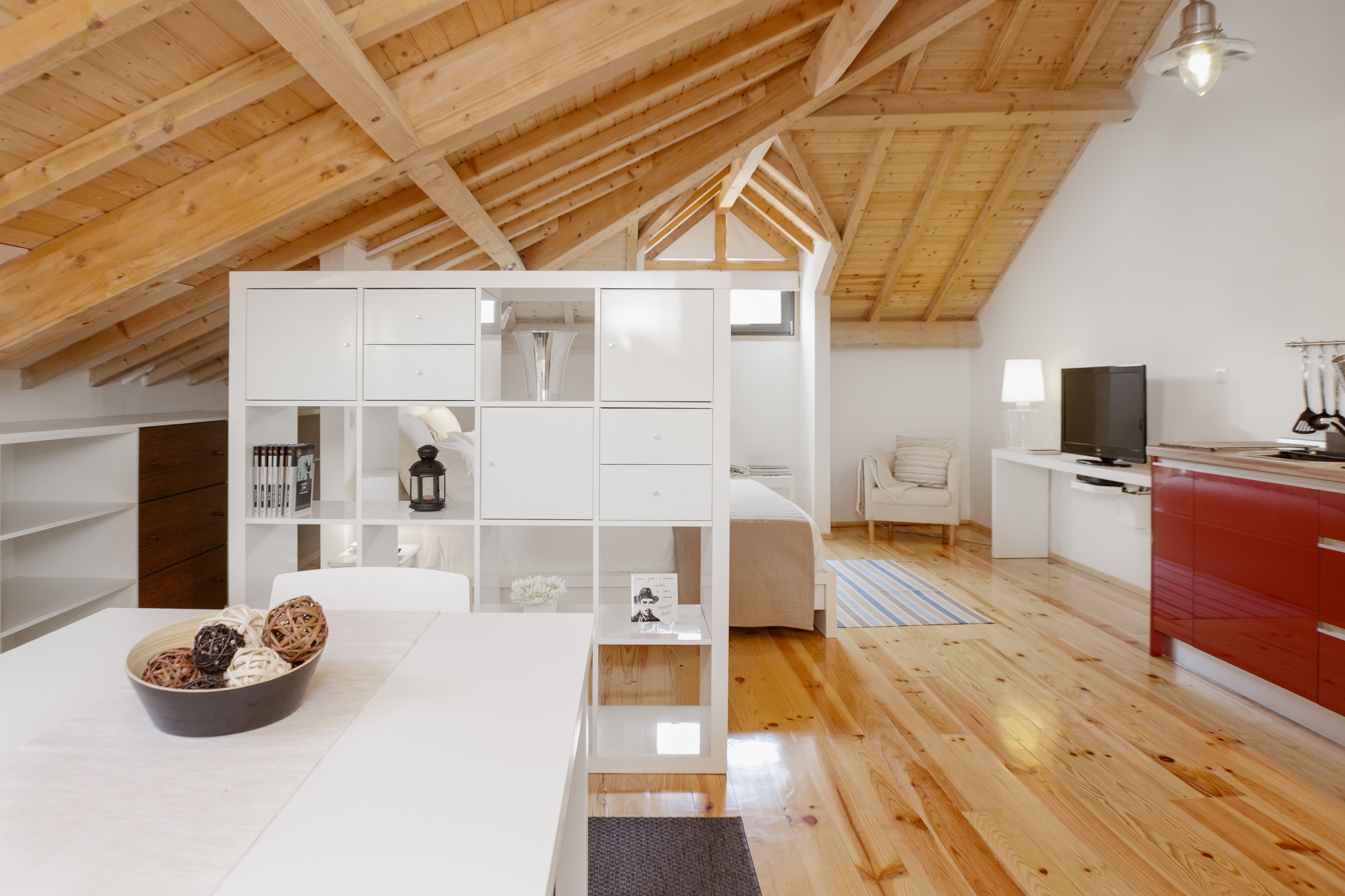 A modern loft bedroom with a sloped ceiling, built-in white shelving, a bed, a desk, and a red kitchen area.