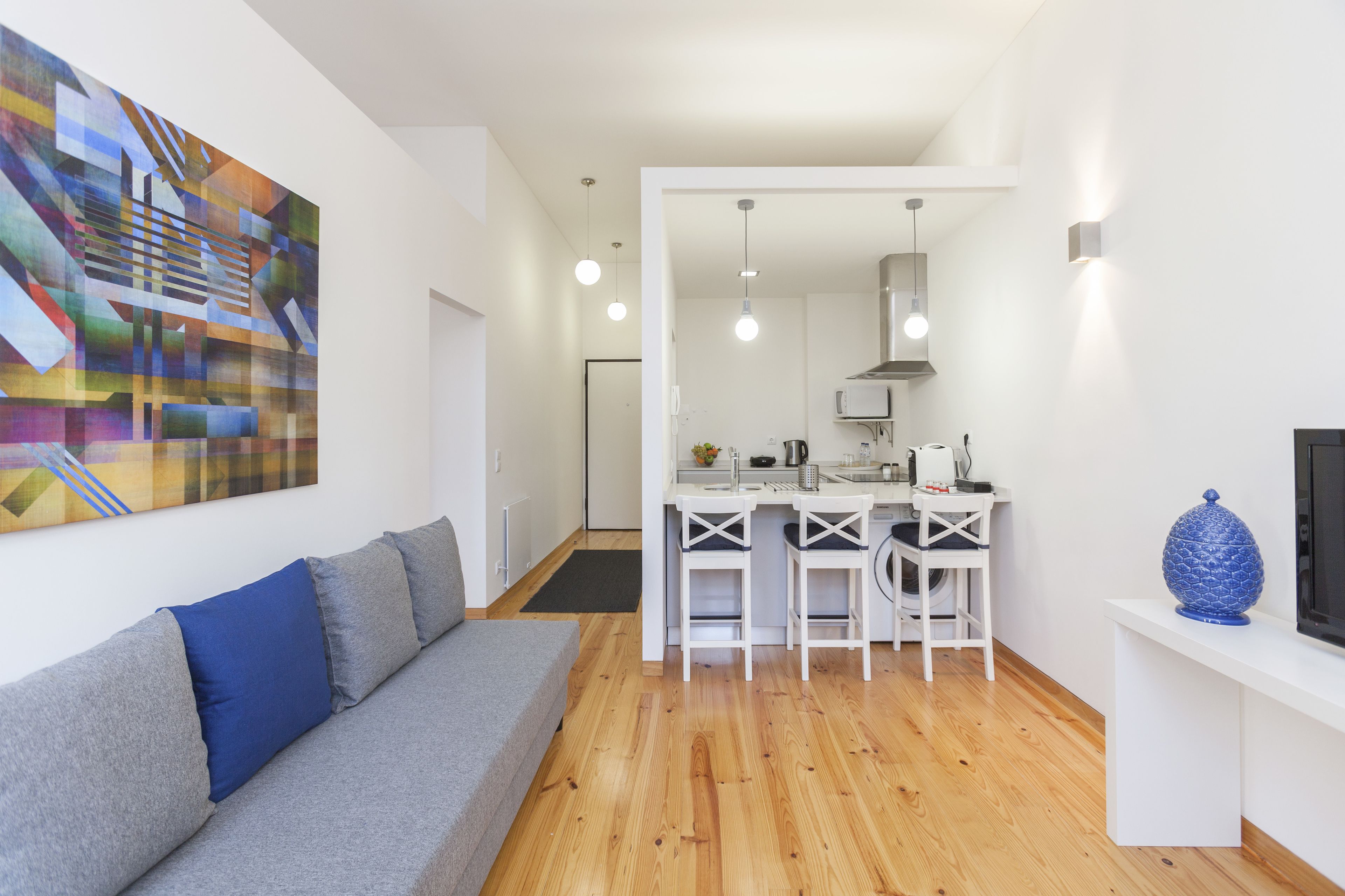 Un appartement moderne avec un canapé gris, une cuisine blanche et un grand tableau abstrait accroché au mur.