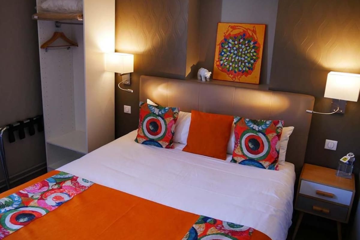 Habitación de hotel con cama, dos lámparas de noche, una mesita de noche, una obra de arte colgada en la pared y un perchero.