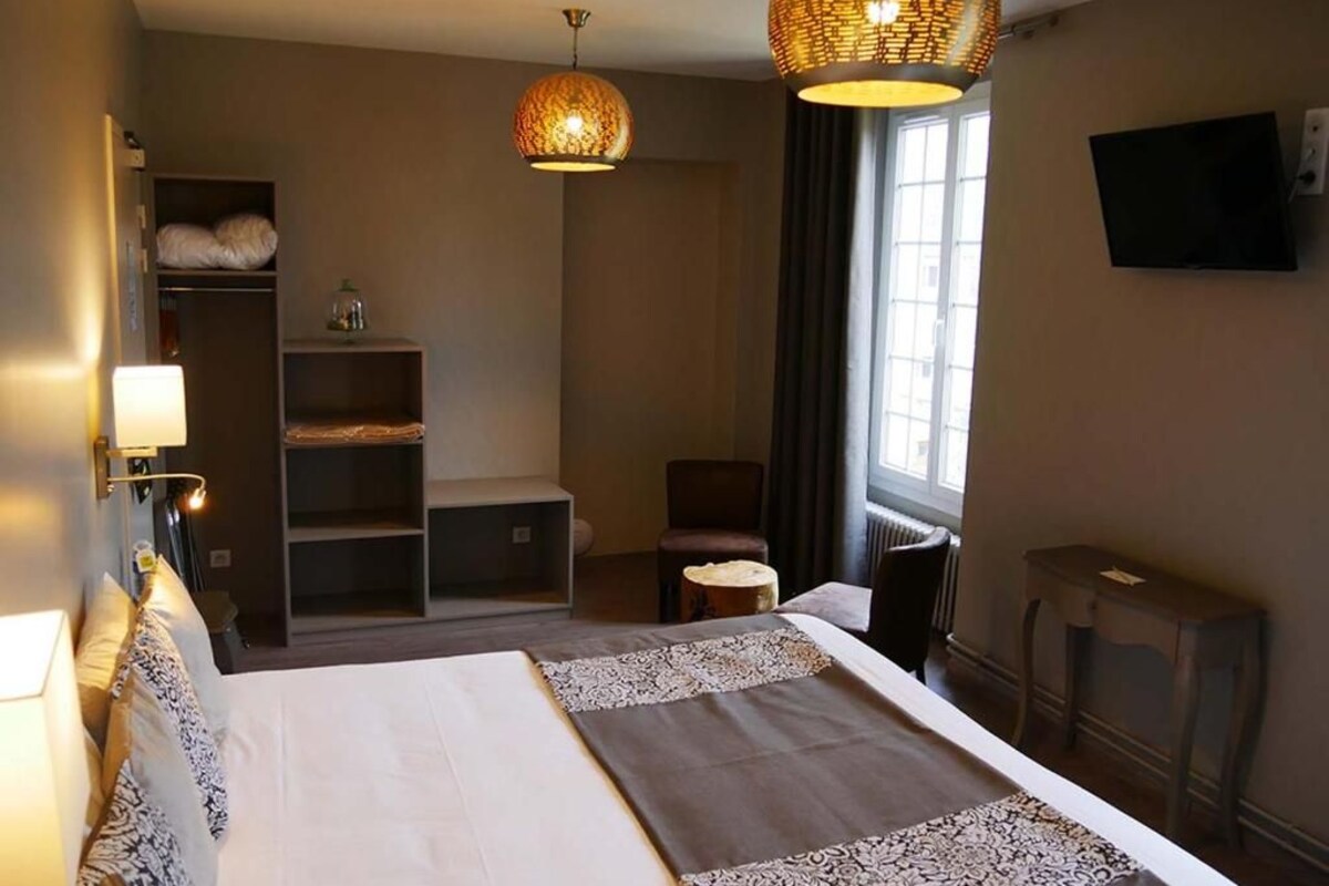 Habitación de hotel con cama, una silla, una mesita, estante con mantas dobladas y dos lámparas colgantes.