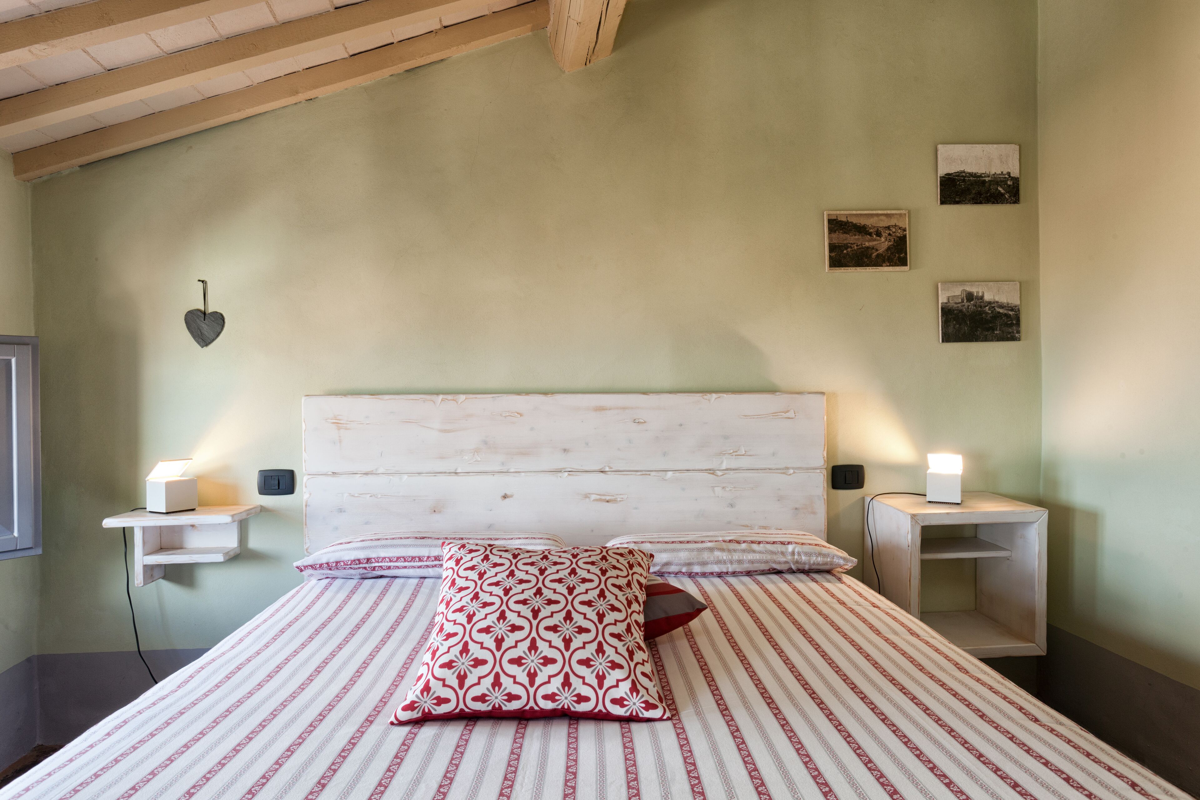 Quarto com cama, duas mesinhas de cabeceira, um criado-mudo, uma luminária na parede e três quadros emoldurados.