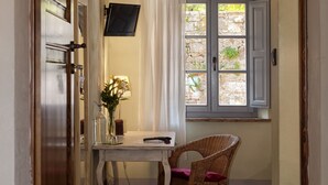 Un couloir étroit avec un plafond en bois, une chaise en osier, une petite table sur laquelle est posé un vase de fleurs et une fenêtre avec des volets.