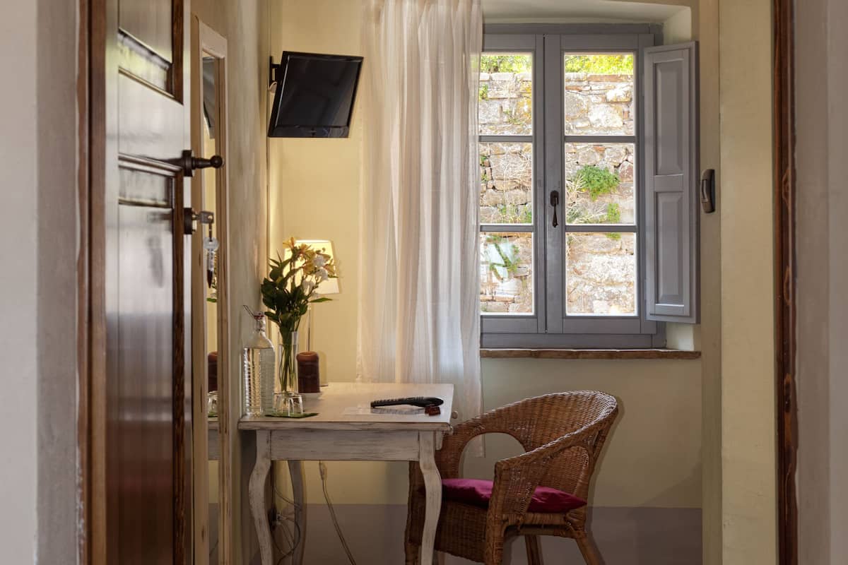 Un couloir étroit avec un plafond en bois, une chaise en osier, une petite table sur laquelle est posé un vase de fleurs et une fenêtre avec des volets.