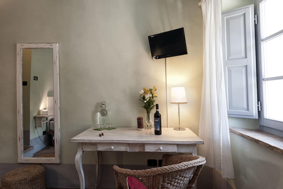Une pièce avec un bureau blanc, un téléviseur à écran plat fixé au mur, une chaise en osier, une lampe et une fenêtre avec des rideaux blancs.
