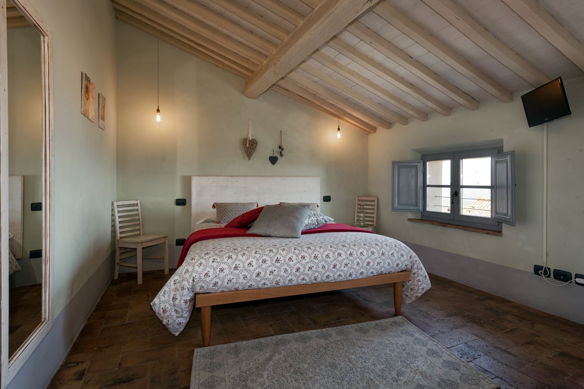 Une chambre avec un lit en bois, une chaise, une fenêtre avec des volets et un téléviseur à écran plat fixé au mur.