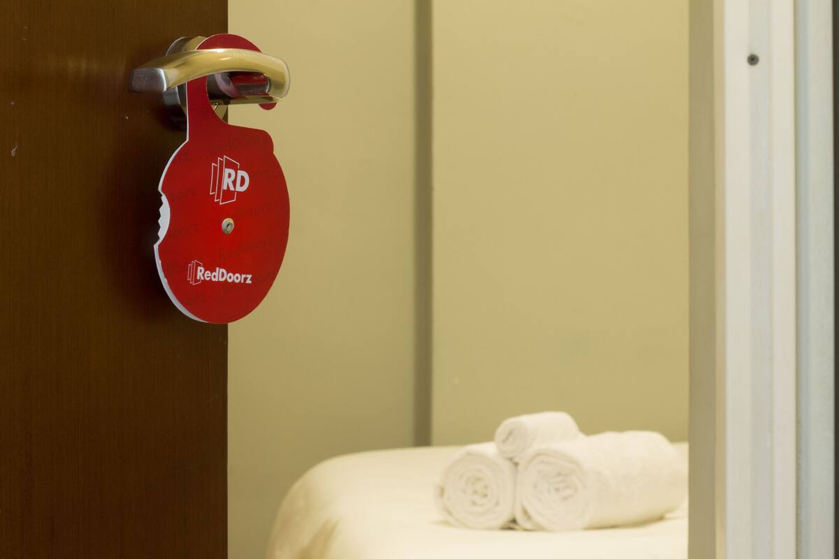 A hotel room with a red door handle, a bed with white linens, and a towel rack with rolled towels.