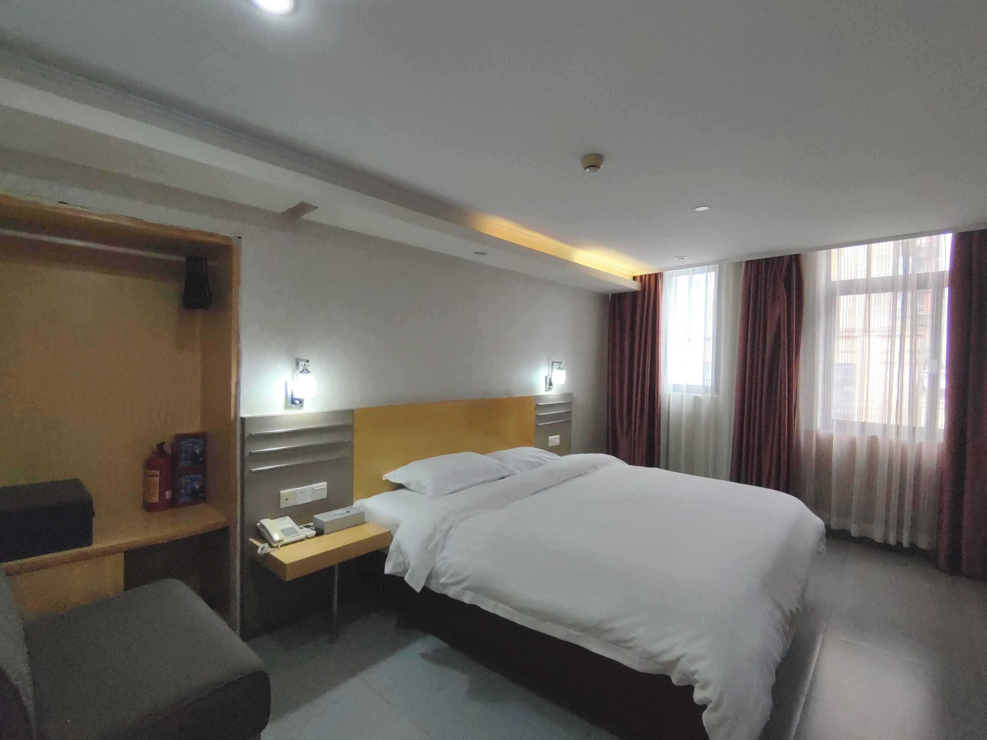 A hotel room with a bed, bedside tables, a desk, a chair, a wardrobe, a window with curtains, and a ceiling light.
