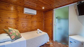 A bedroom with wooden panel walls, a bed with a white bedspread and a floral pillow, a white air conditioning unit, a television, and a refrigerator. - Fazenda Pousada das Palmeiras (Brotas)