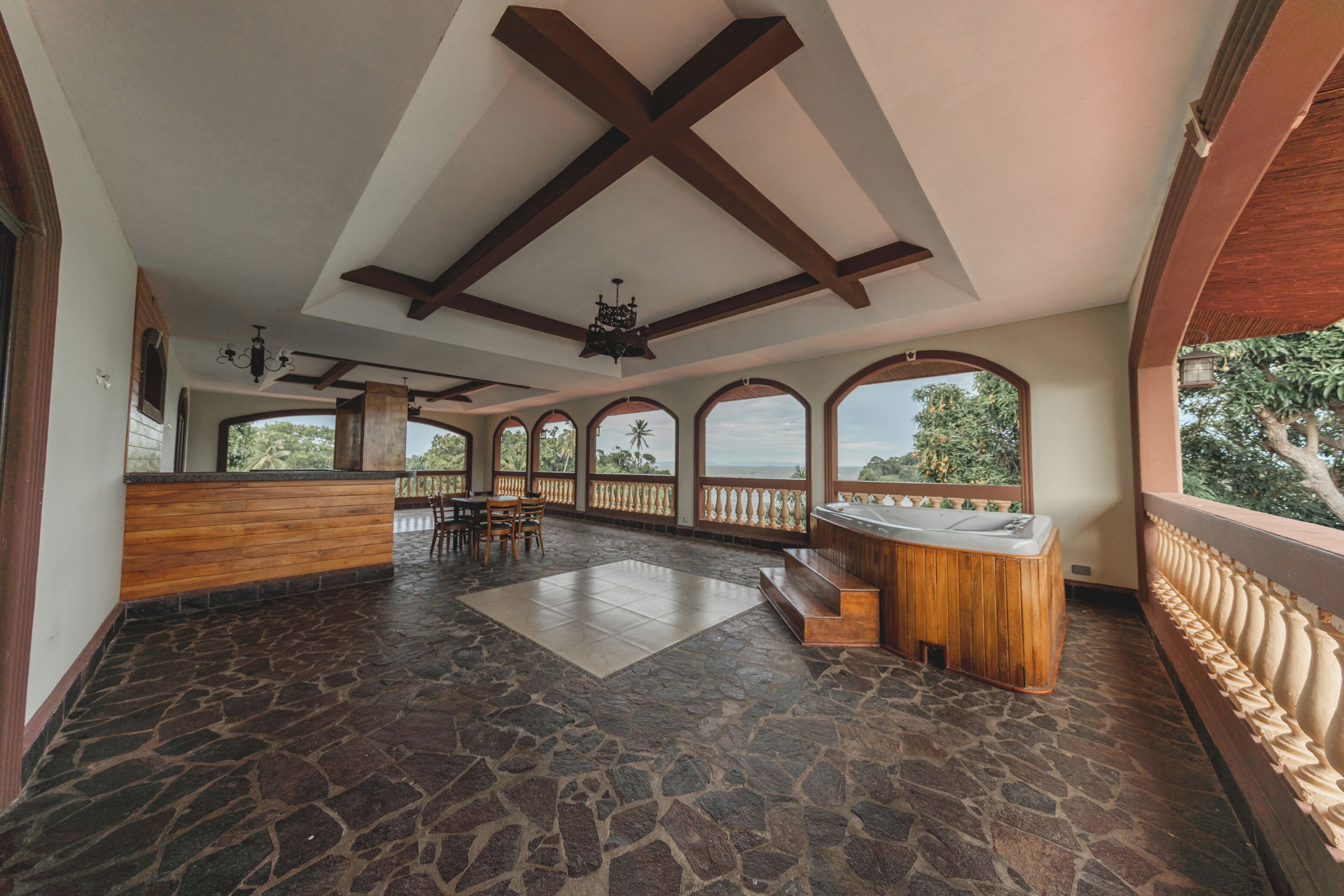 A spacious room with a stone floor, wooden reception desk, and large arched windows offering a view of the sea.