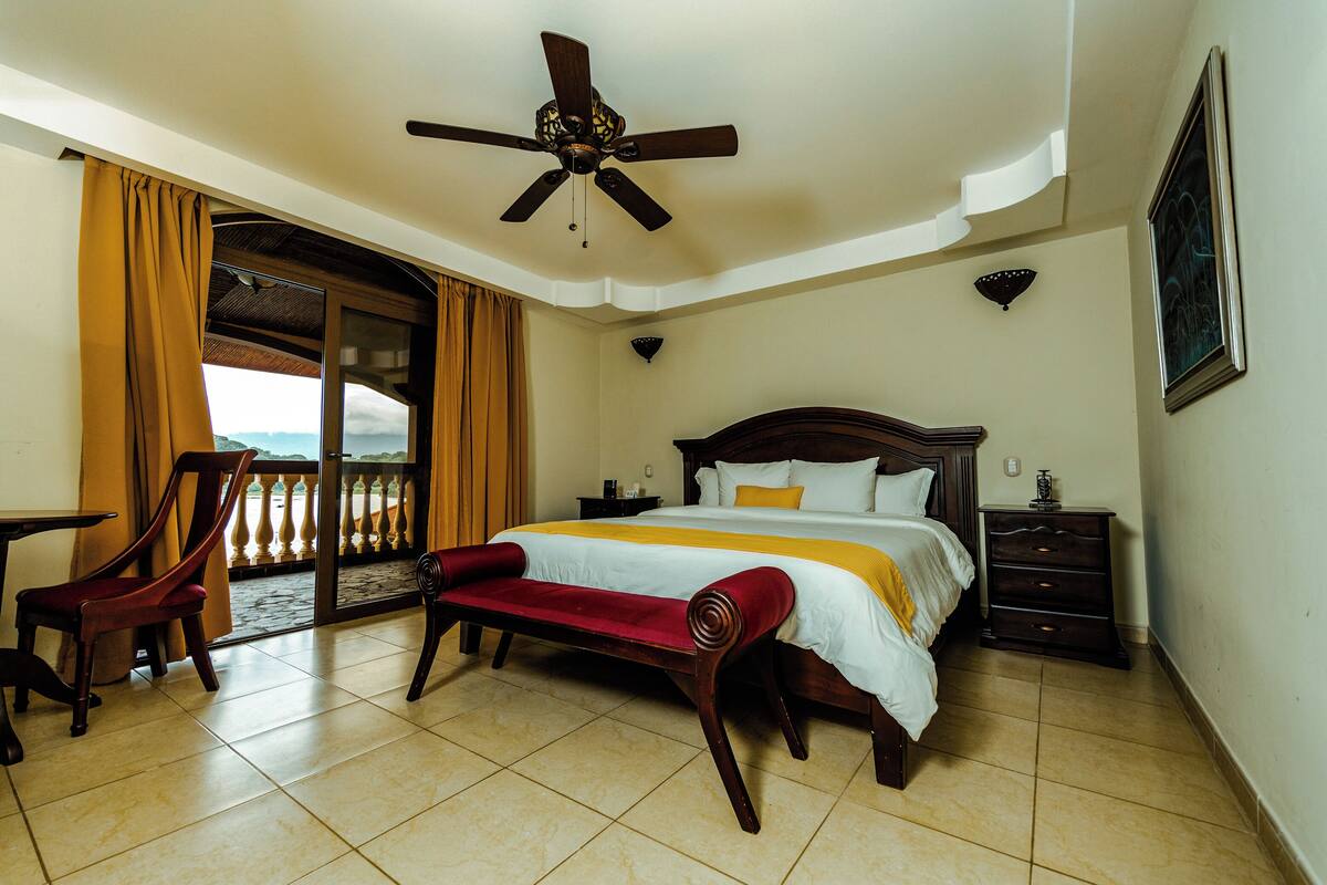 Habitación de hotel con una cama, un banco de madera, un ventilador de techo, una mesita de noche y vistas al exterior.