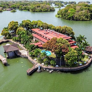 Vista aérea de un resort con piscina, un puente y un edificio de techo rojo, rodeado de exuberante vegetación y agua.