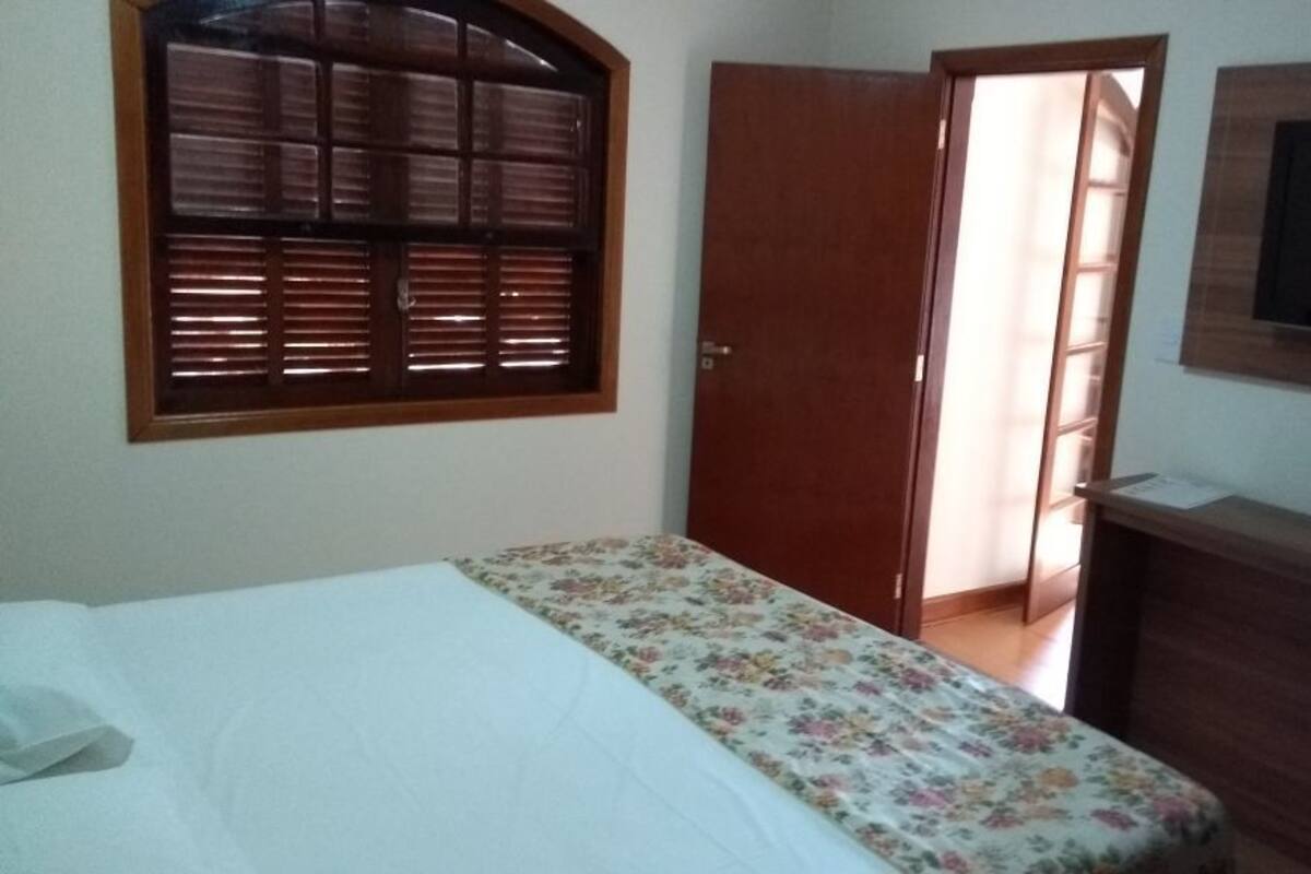 Un dormitorio con una cama, una puerta de madera, un televisor montado en la pared y una ventana con persianas.