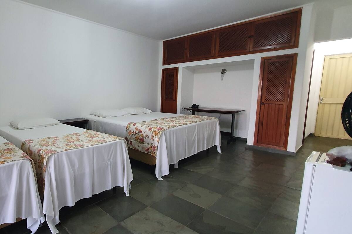 Una habitación con tres camas individuales, cada una con cubrecama floral y almohadas blancas. Se observa un armario de madera sobre las camas y una pequeña mesa con una silla.