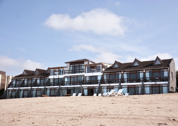 A large hotel with multiple floors, a beach in front, and several lounge chairs.