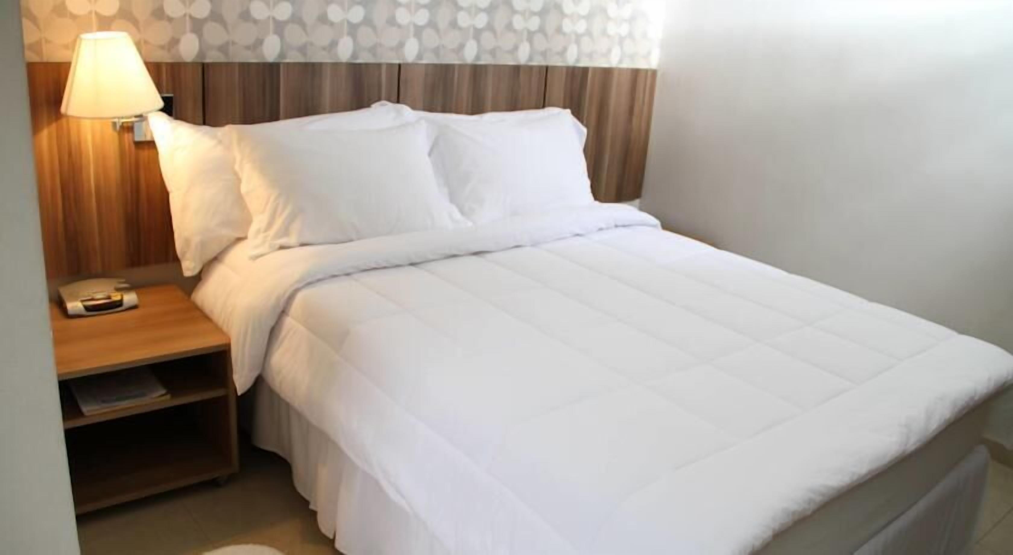 Una cama bien hecha con ropa de cama blanca, una mesita de noche de madera con una lámpara y un papel tapiz estampado.