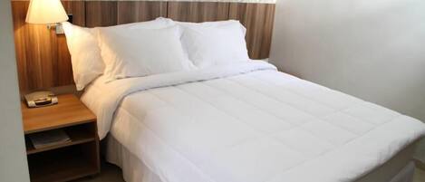 Una cama bien hecha con ropa de cama blanca, una mesita de noche de madera con una lámpara y un papel tapiz estampado.