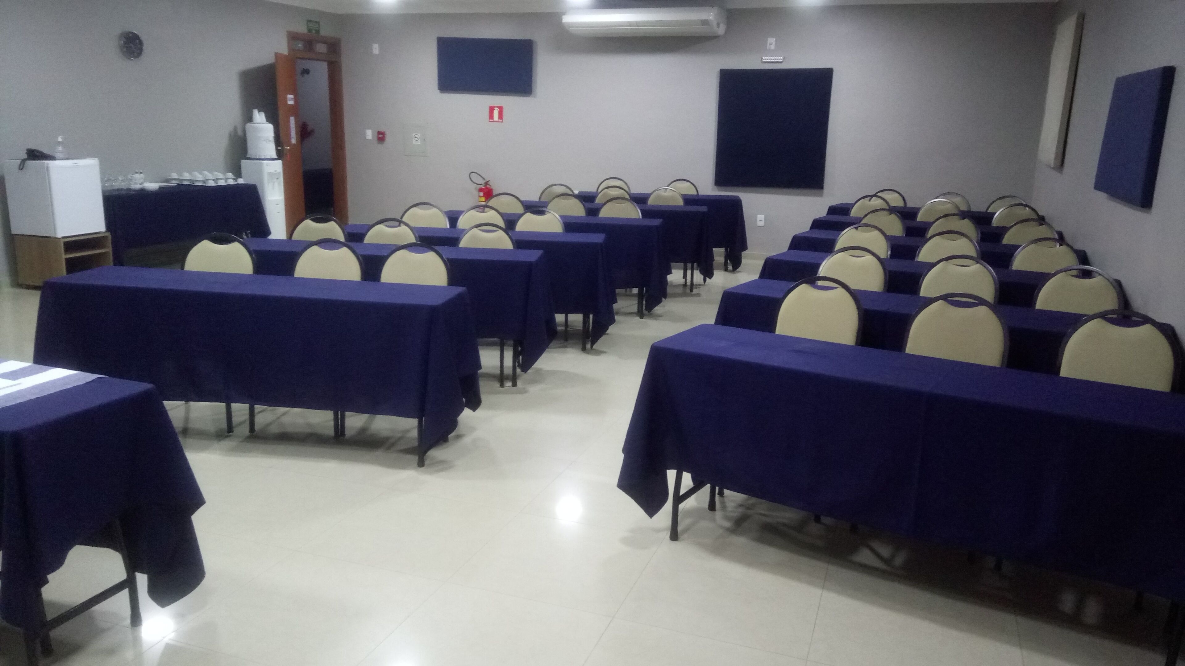 Un salón de conferencias con filas de mesas y sillas, una pizarra y un dispensador de agua.
