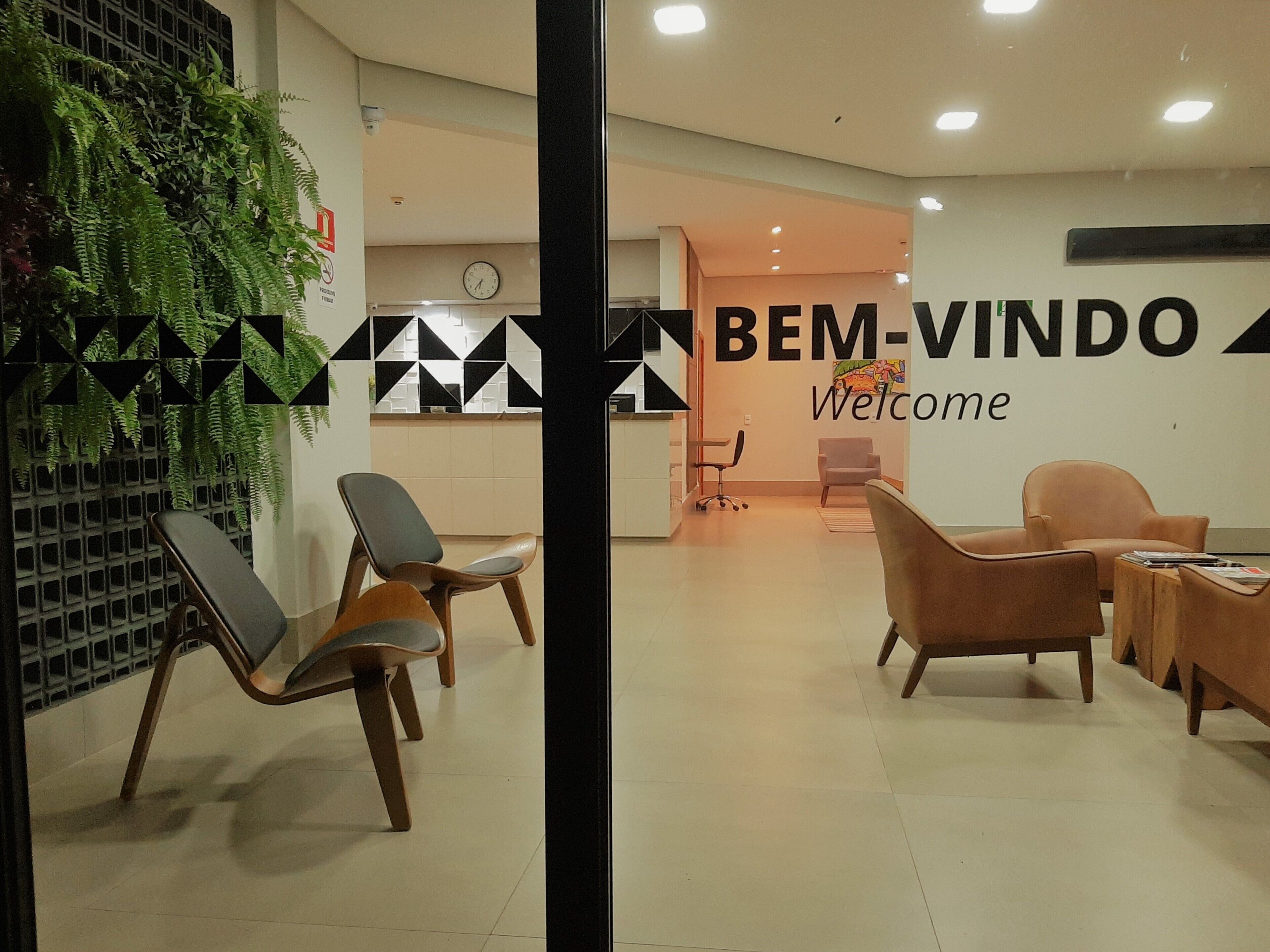 A reception area with a 'BEM-VINDO Welcome' sign, several chairs, a clock, and a plant.
