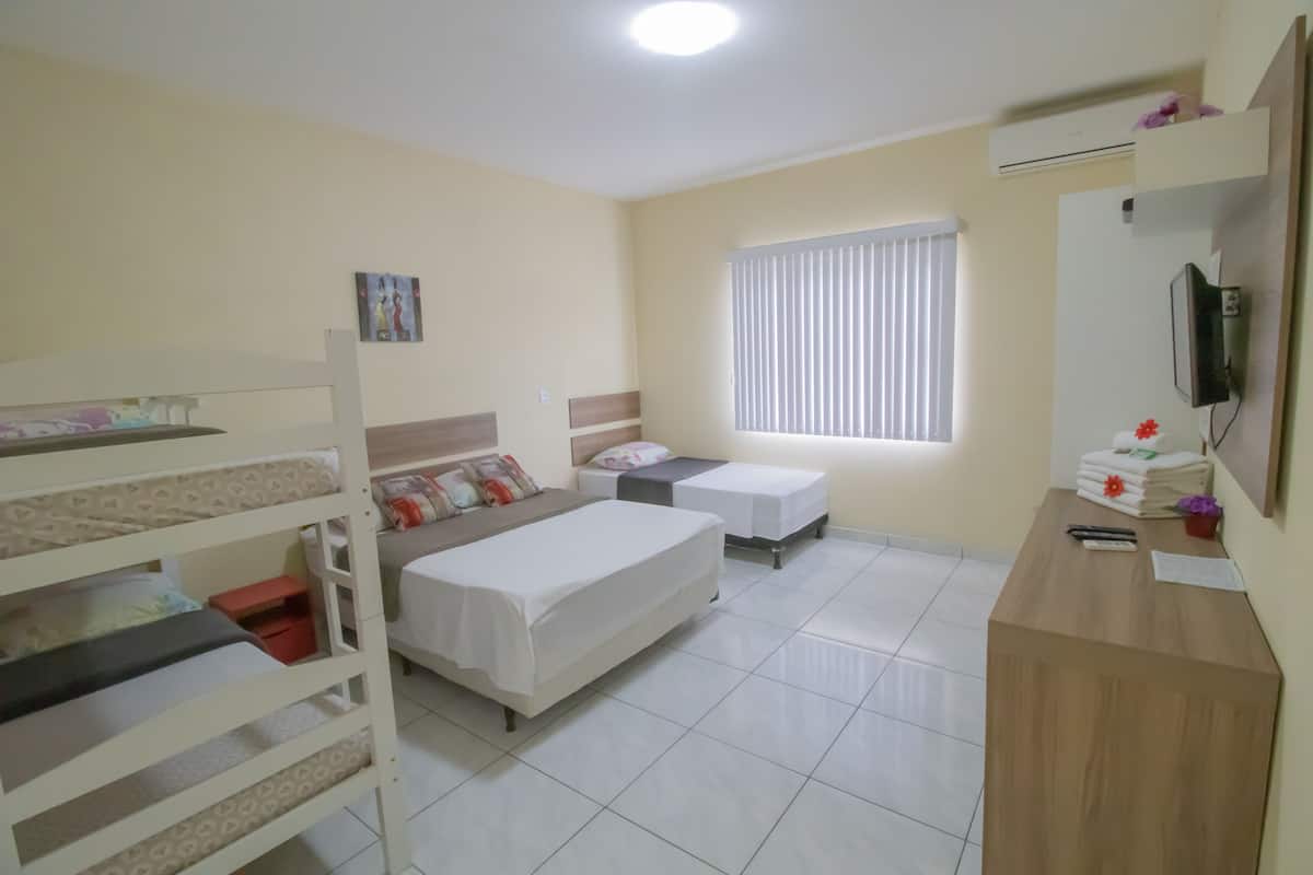 A hotel room with bunk beds, a single bed, a desk with a mirror, and a window with blinds.