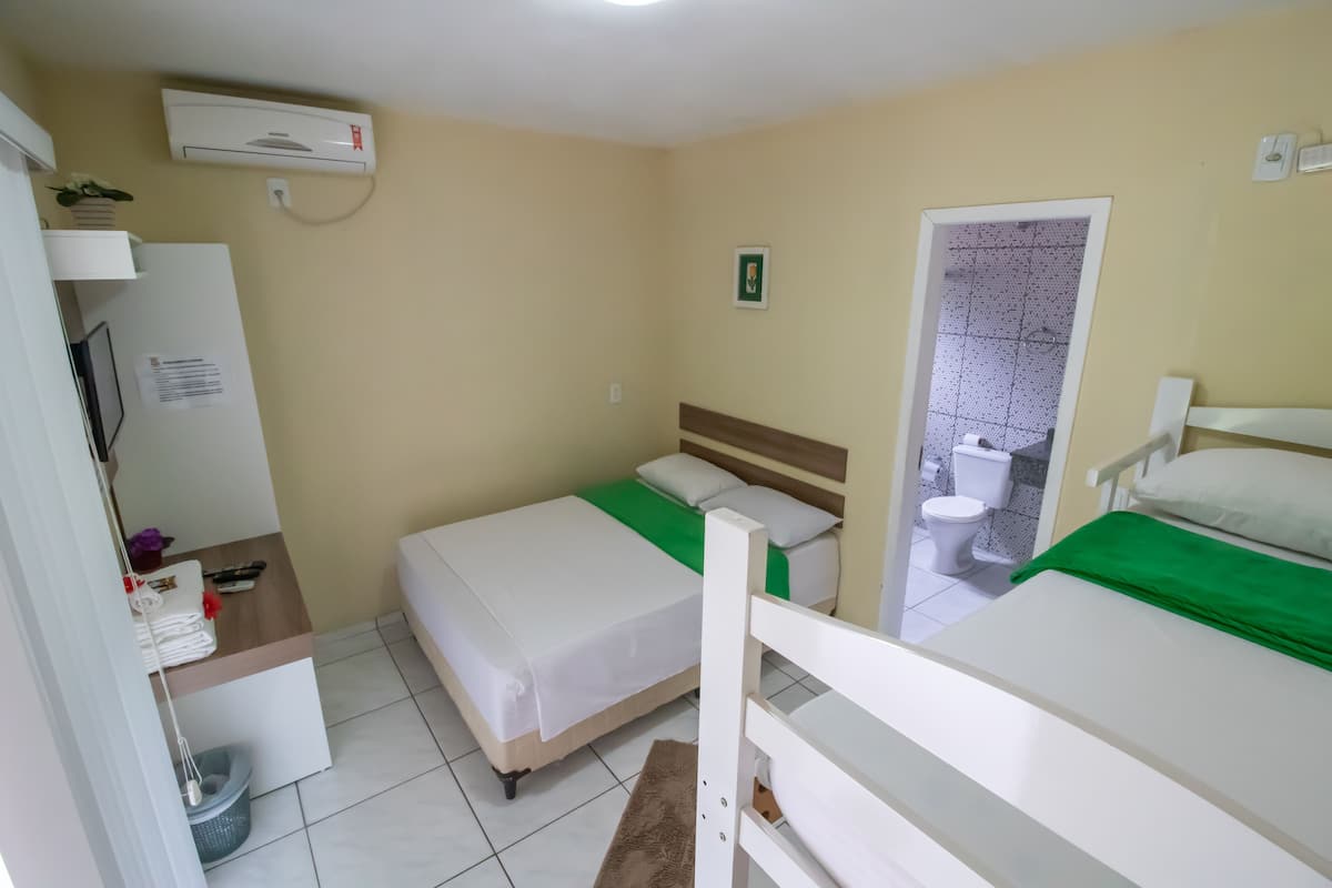 A small, clean room with two beds, a toilet, and a sink. There is a white air conditioning unit on the wall and a small shelf with a plant.