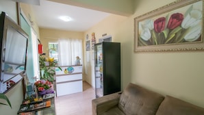 A living room with a sofa, a TV, a refrigerator, and a kitchen area with a counter and a plant.