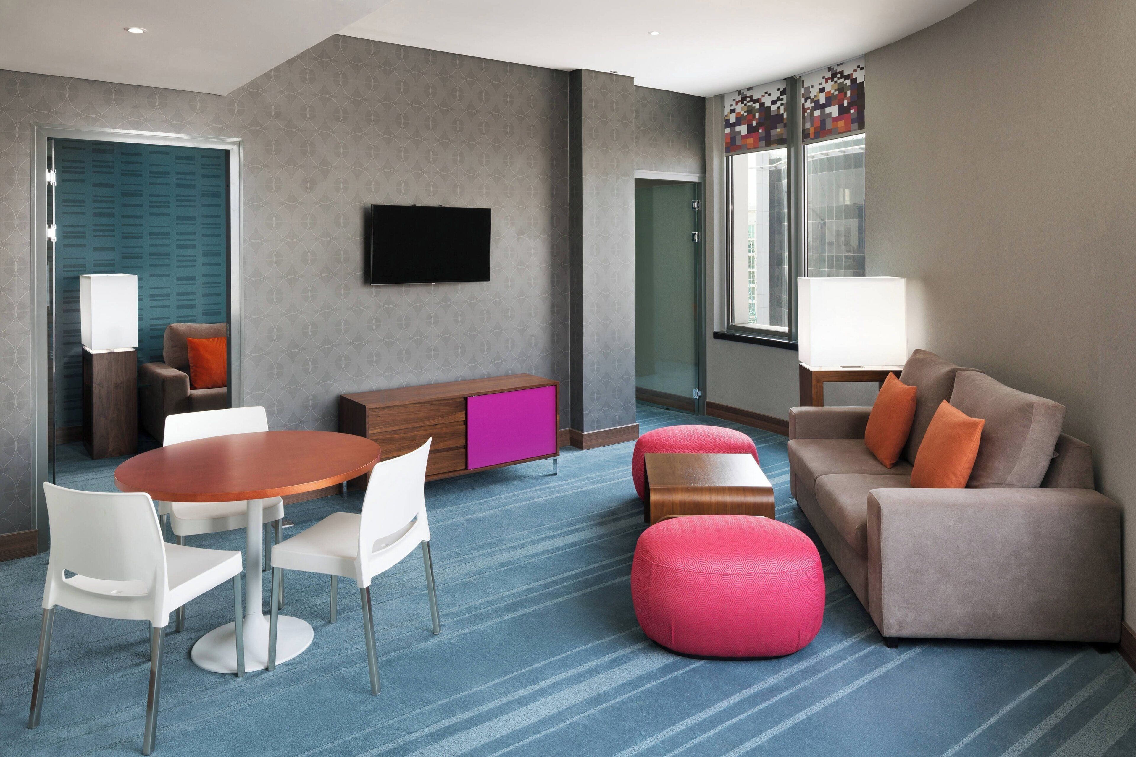 A modern hotel room with a round dining table, white chairs, a sofa, and a pink ottoman.