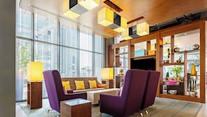A modern lobby with purple armchairs, a sofa, and a coffee table. There are large windows, a wooden bookshelf, and a TV. - Aloft by Marriott Riyadh (Riyadh)