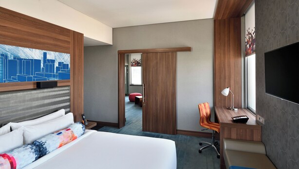 A hotel room with a large bed, a desk with a chair, a TV, and a cityscape artwork on the wall.