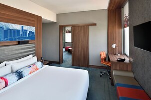 A hotel room with a large bed, a desk with a chair, a TV, and a cityscape artwork on the wall.