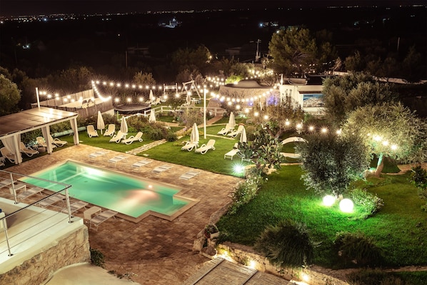 A pool area with lounge chairs and umbrellas, surrounded by a well-lit garden and a view of the city at night.