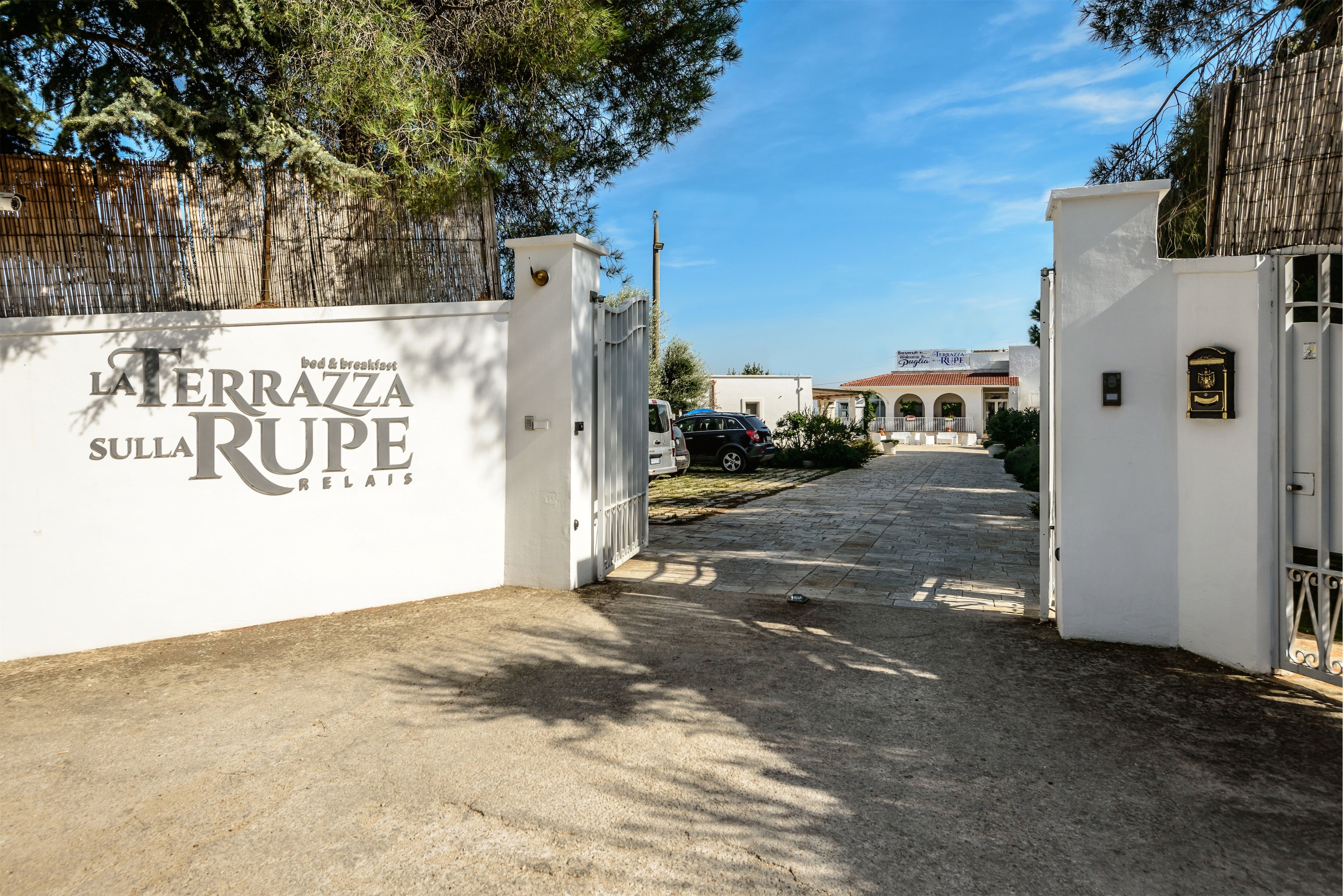 Een toegangspoort naar een landgoed genaamd 'Terrazza Sulla Rupe Relais' met een verharde oprijlaan die leidt naar een gebouw met een rode dakbedekking.