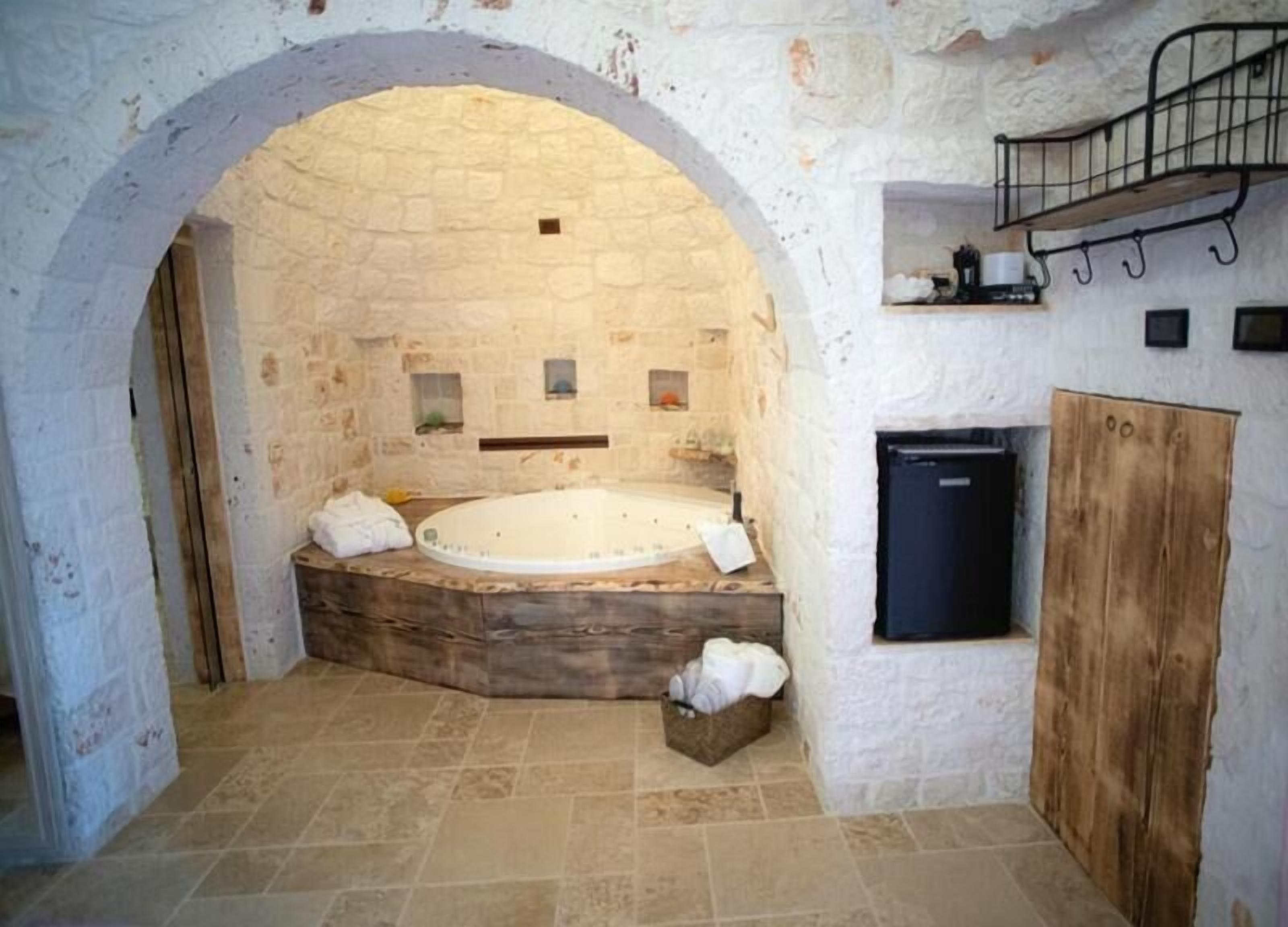Una habitación con paredes de piedra, una bañera de madera, un refrigerador pequeño y un banco de madera.
