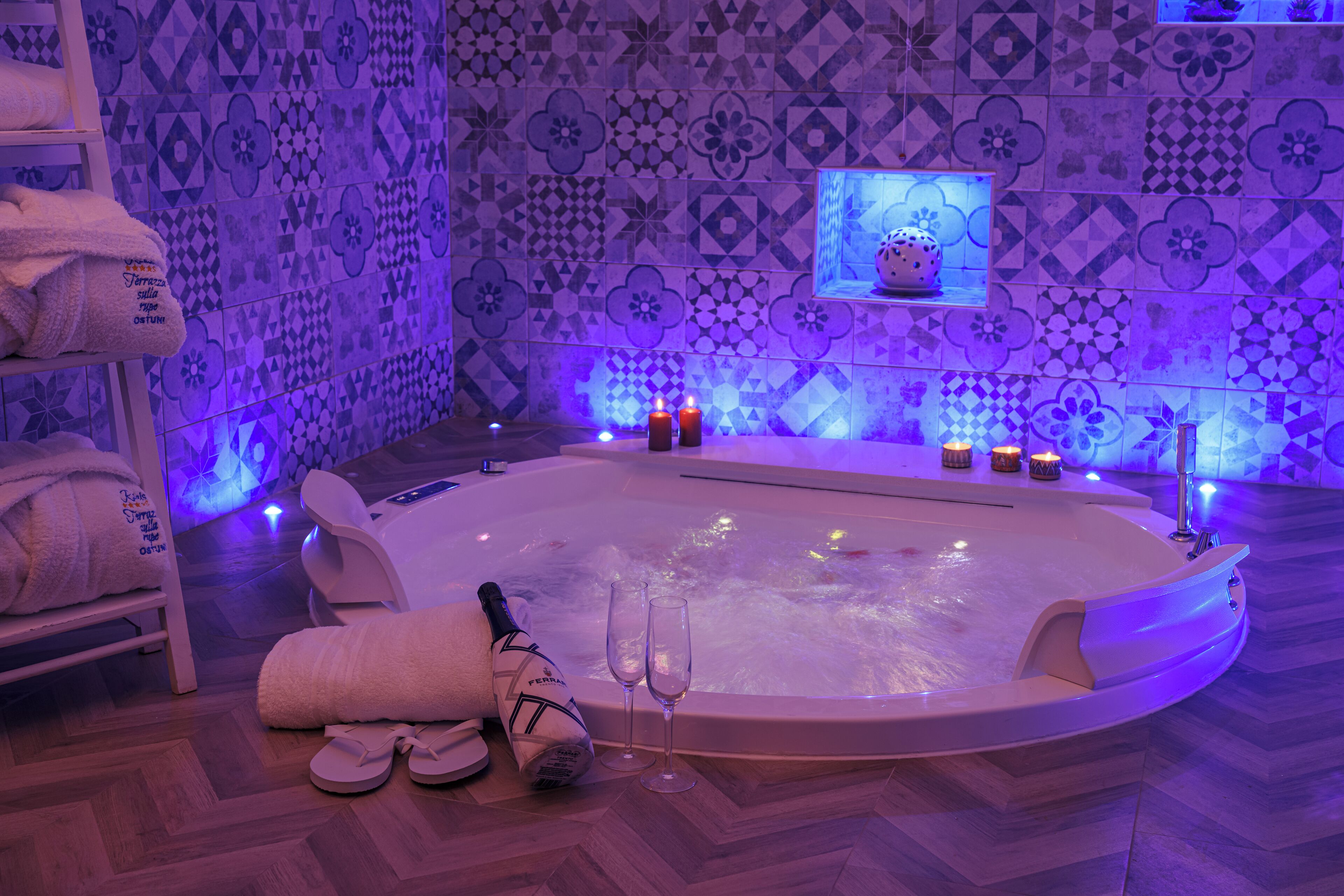 A spa room with a large bathtub, a shelf with towels, a bottle of champagne, and two glasses.