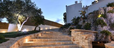 Una escalera de piedra que sube hacia un edificio de fachada blanca y un árbol al fondo.