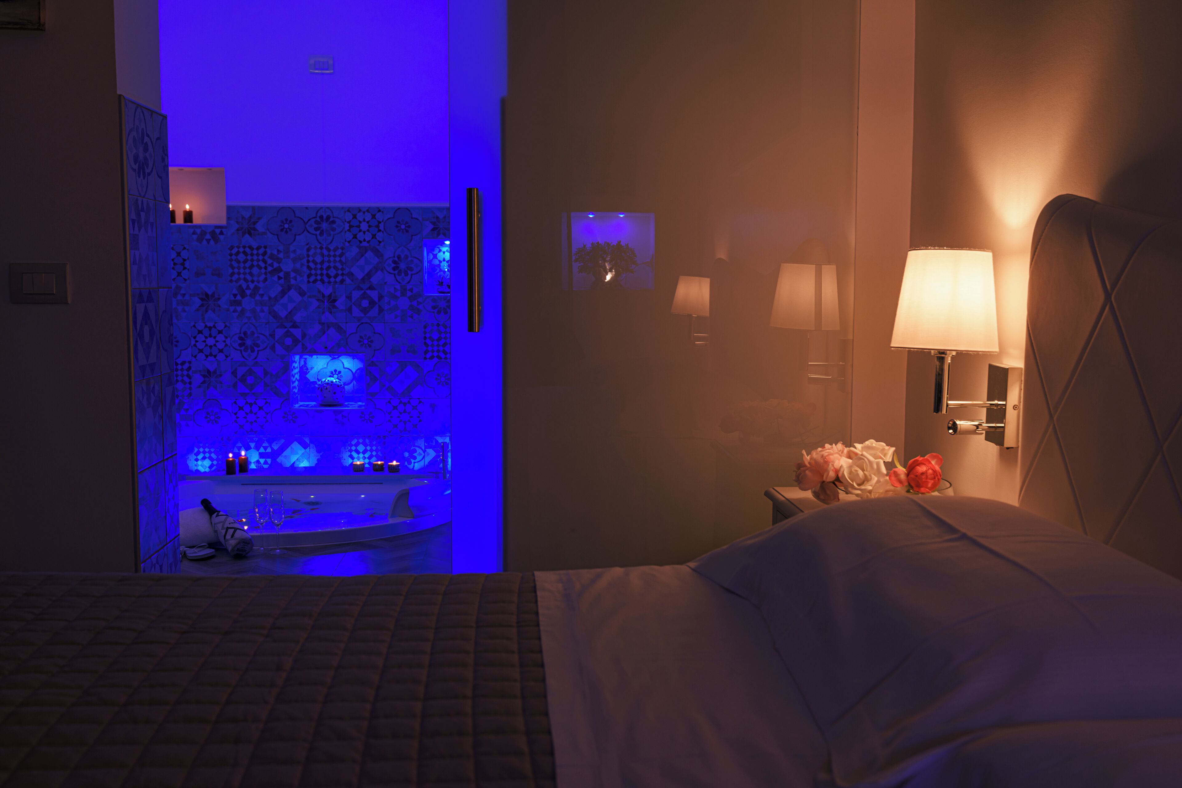 Quarto de hotel com cama, mesa de cabeceira com abajur, televisão na parede e banheiro com área de chuveiro iluminada em azul.