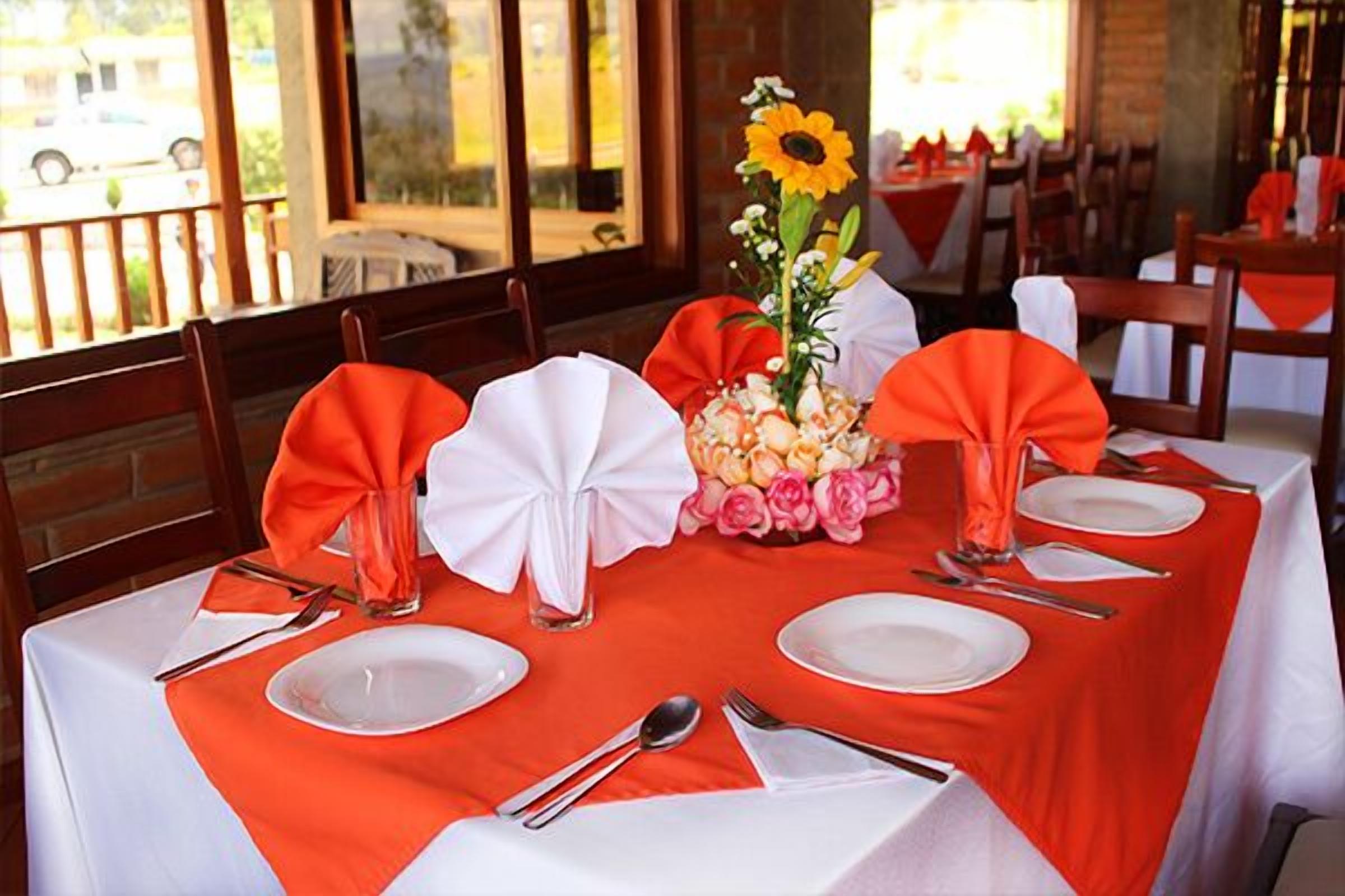 A dining table set with white plates, silverware, and orange napkins. A vase with flowers and a sunflower is on the table.