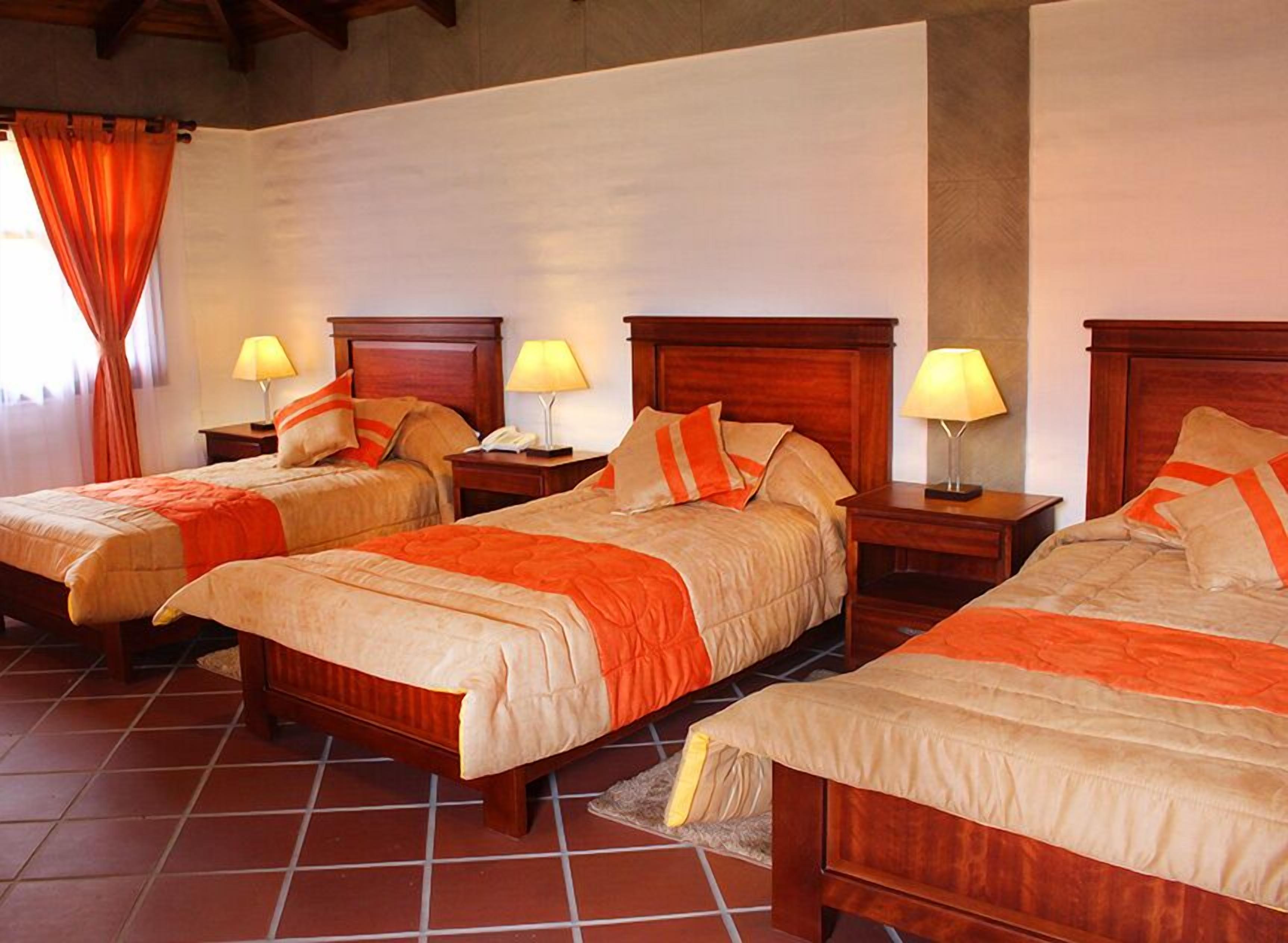 Habitación de hotel con tres camas individuales, cada una con cabecero de madera, mesita de noche y lámpara. El piso es de baldosas y hay una ventana con cortinas.