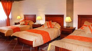 Habitación de hotel con tres camas individuales, cada una con cabecero de madera, mesita de noche y lámpara. El piso es de baldosas y hay una ventana con cortinas.