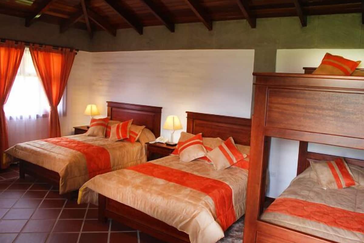A hotel room with two beds, each with a wooden headboard and red bedding. There are two bedside tables with lamps. The room has a tiled floor and a wooden ceiling.