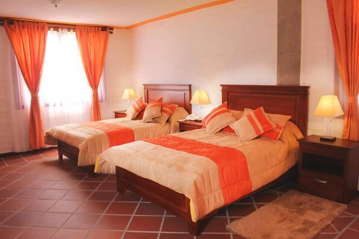 A hotel room with two beds, wooden headboards, and orange curtains.