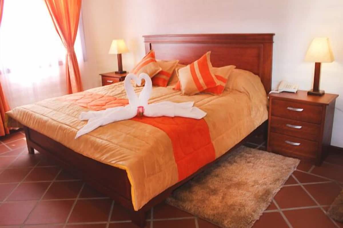 A neatly made bed with a red and beige bedspread, two pillows, and a decorative swan made of towels. There are two bedside tables with lamps, a wooden headboard, and a tiled floor.