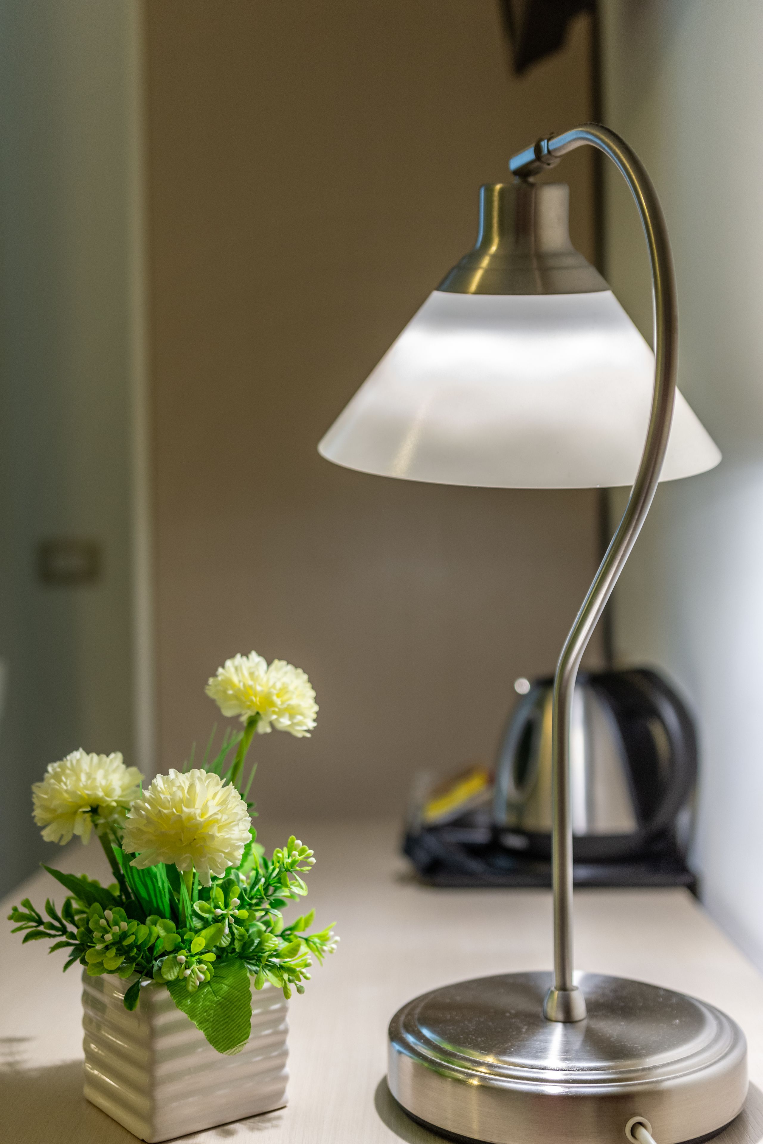 A desk lamp with a white shade and a metallic base, positioned next to a white vase with green foliage and white flowers.