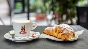 A cup of coffee and a croissant on a table with a blurred outdoor background. - B&B Cairoli (Messina)