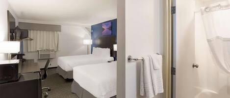 Habitación de hotel con dos camas, un escritorio con computadora, una silla, una lámpara de escritorio, un cuadro en la pared y un baño con cortina de ducha.