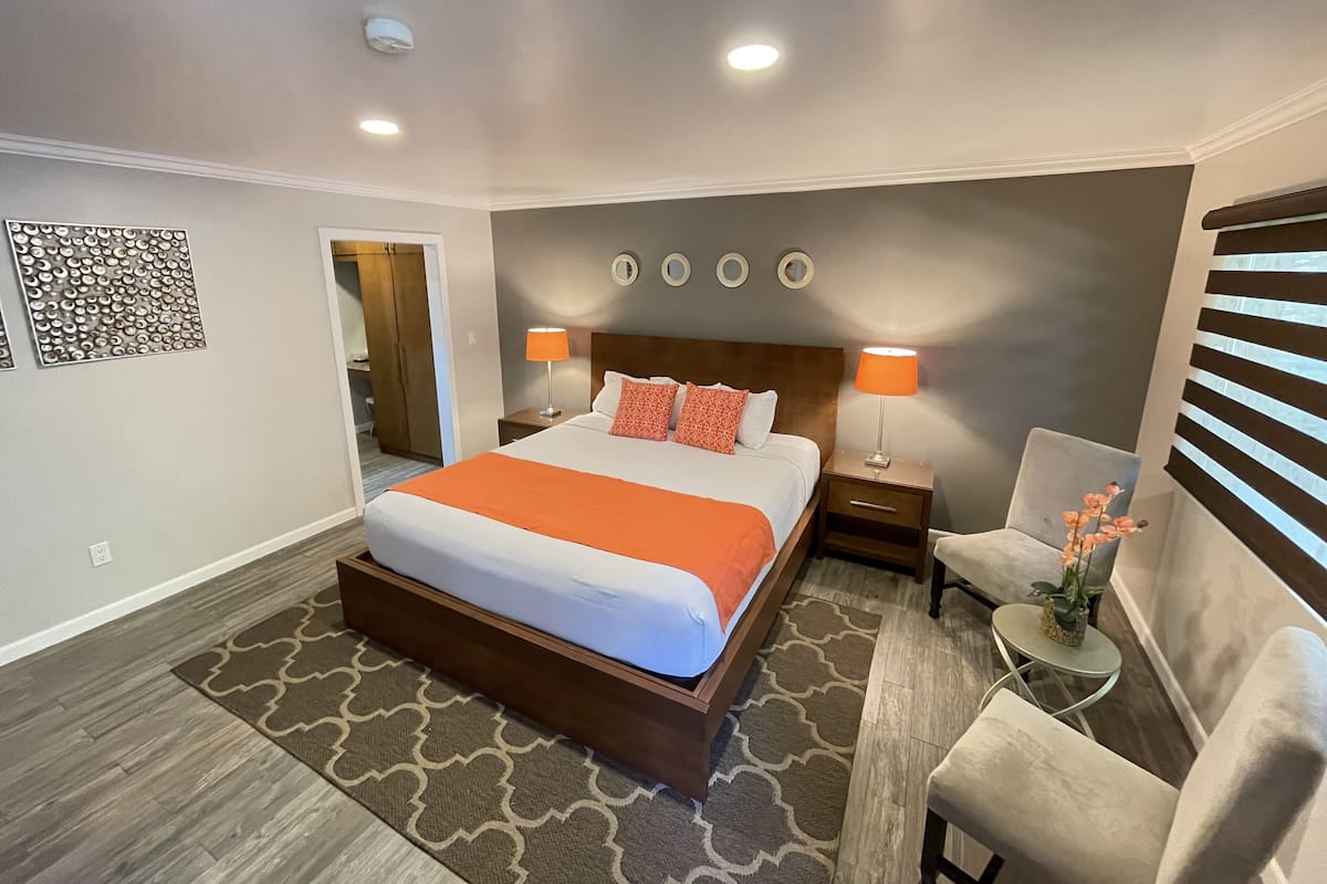 A hotel room with a large bed, two bedside tables with lamps, a chair, a small table with a plant, and a decorative wall piece.