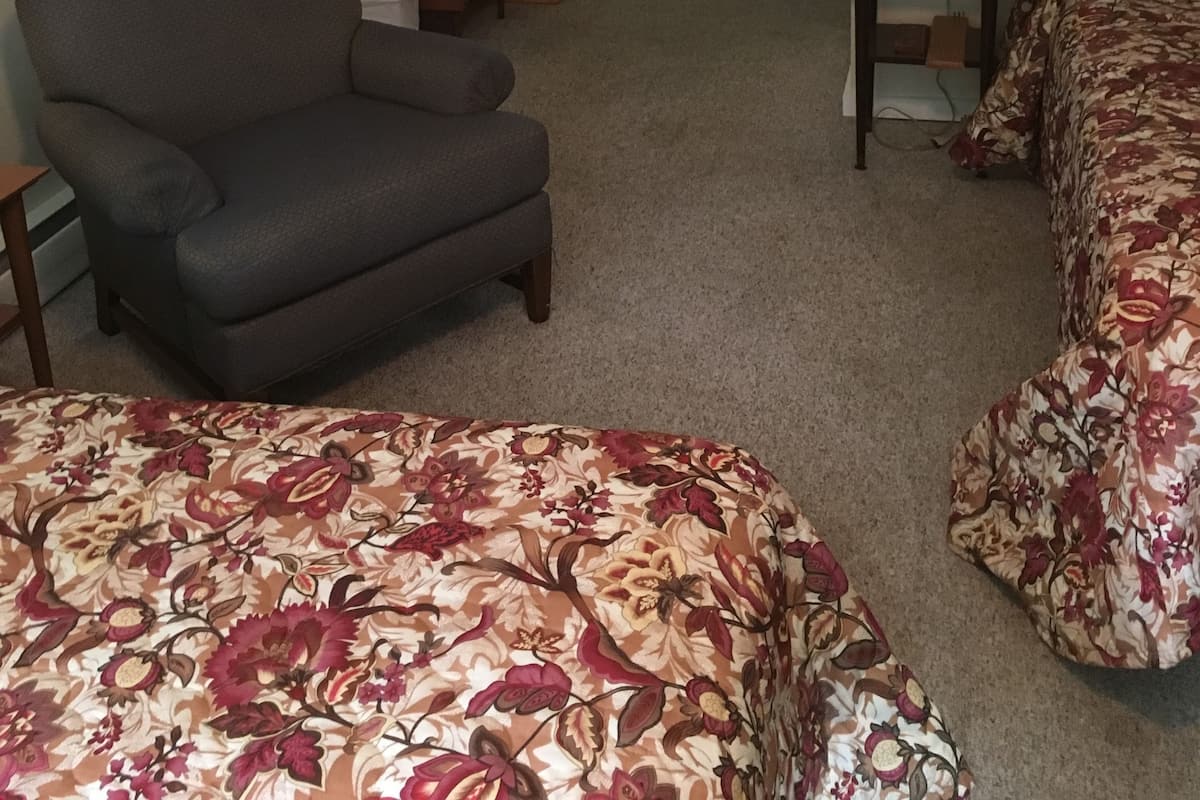 A room with a grey armchair, a bed with floral bedding, a small table with a lamp, a dresser, and a television.