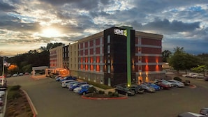 Un hotel de diseño moderno, con un llamativo letrero 'HOME 2'. Tiene varios pisos con diferentes tipos de ventanas y un estacionamiento con varios automóviles.