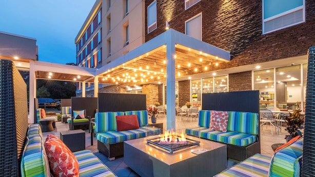 Un espacio al aire libre con cojines de rayas coloridas, una chimenea central y un porche cubierto con luces colgantes.