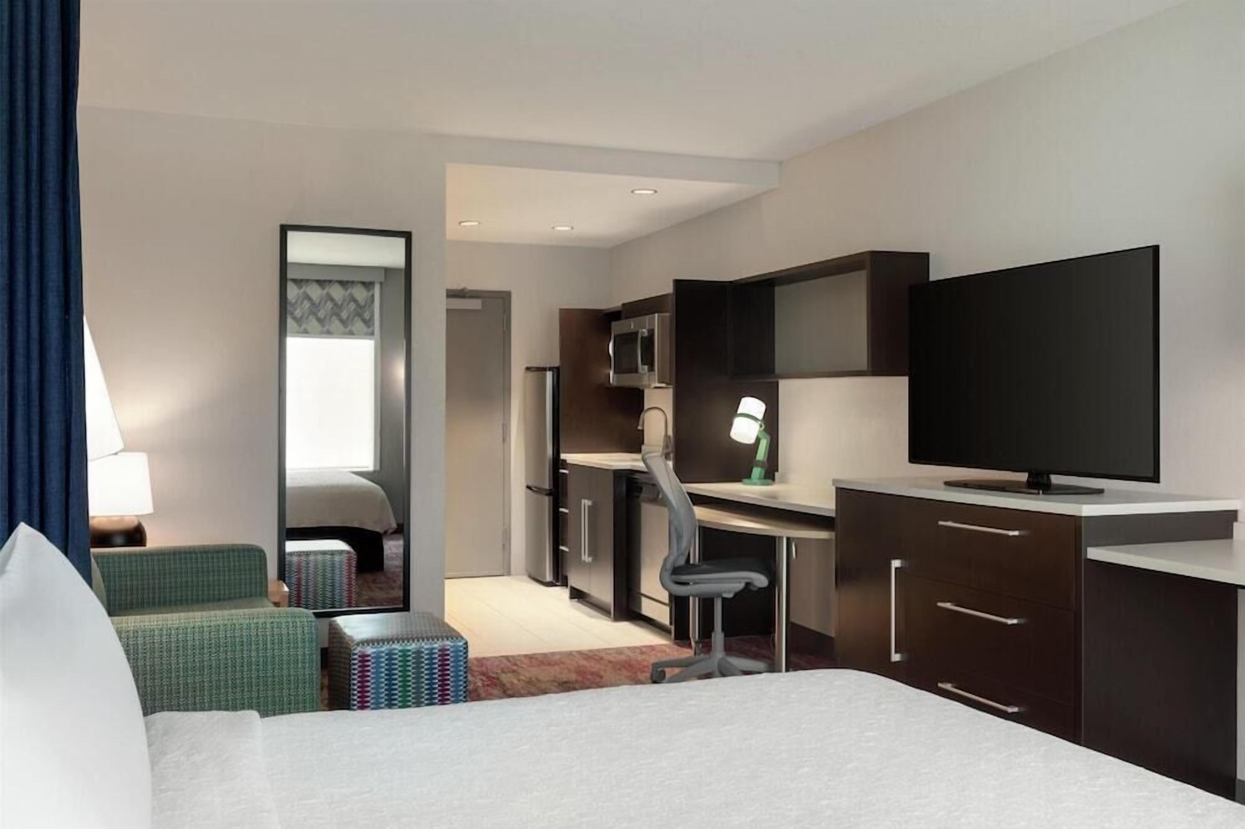 Quarto de hotel com cama, escrivaninha, cadeira, televisão, espelho e cozinha compacta.