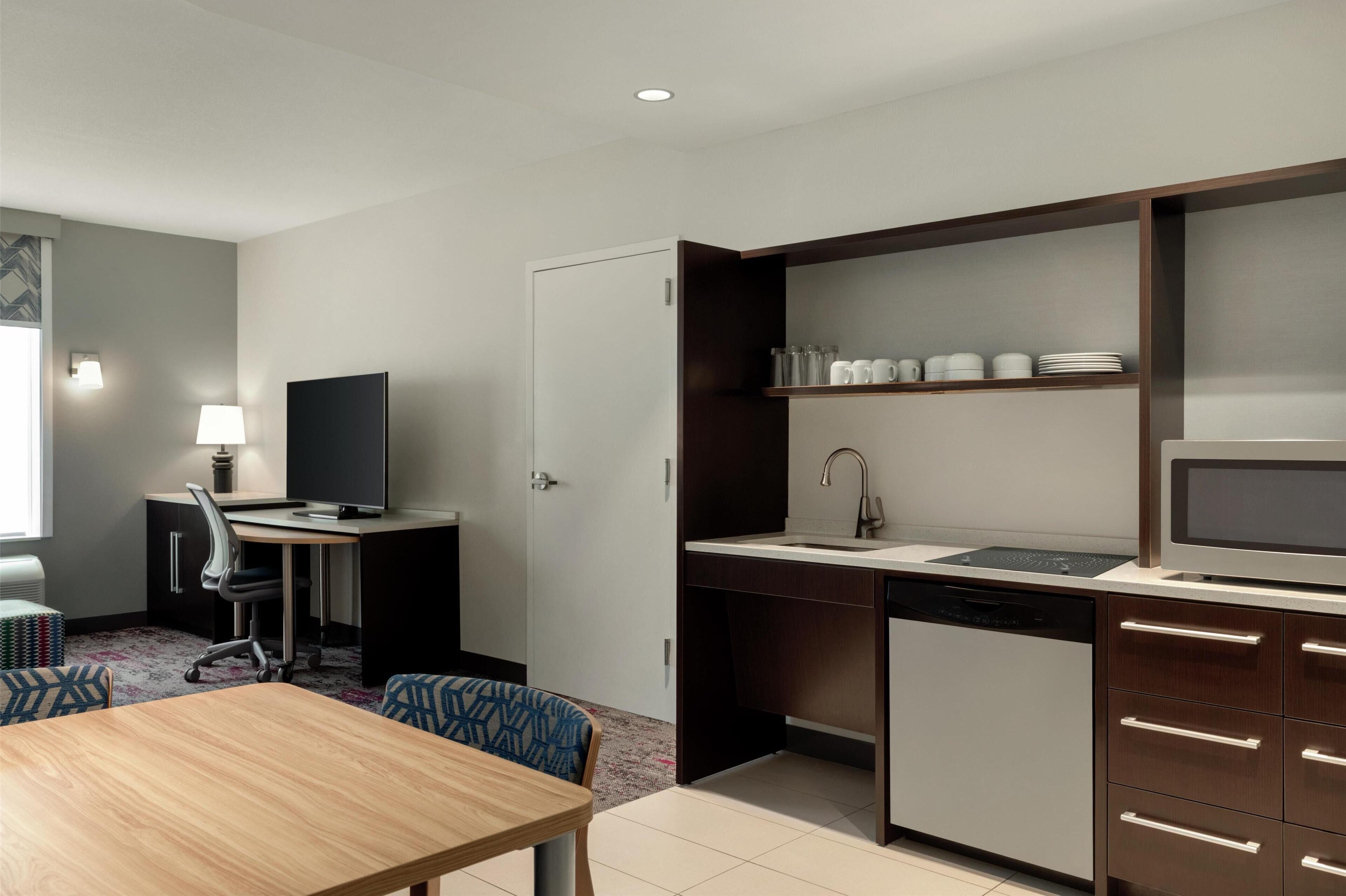 A compact hotel room with a kitchenette, a dining area, a desk with a chair, and a television.